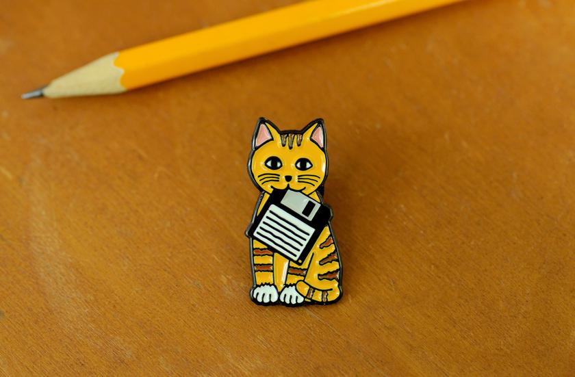 Enamel pin with smiling orange tabby kitty sitting holding a floppy disk in its mouth.