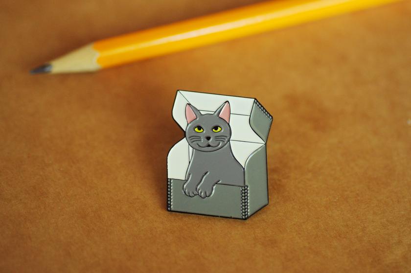 Enamel pin with smiling gray kitty sitting in an archival box with the lid open.