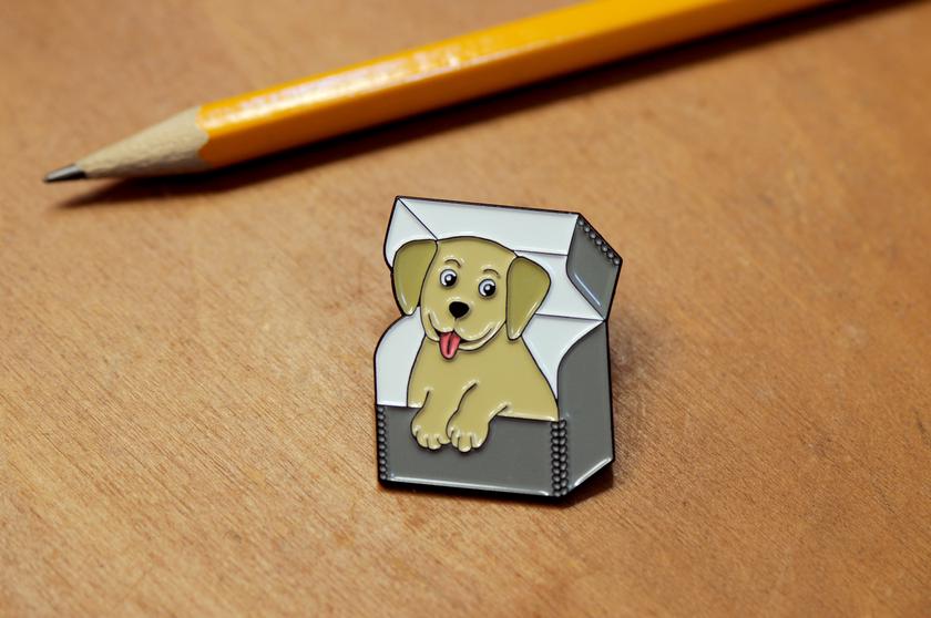 Enamel pin with smiling doggy sitting in an archival box with the lid open.